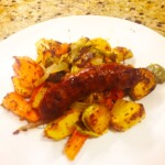 Brats with roasted vegetables on a white plate on a counter.