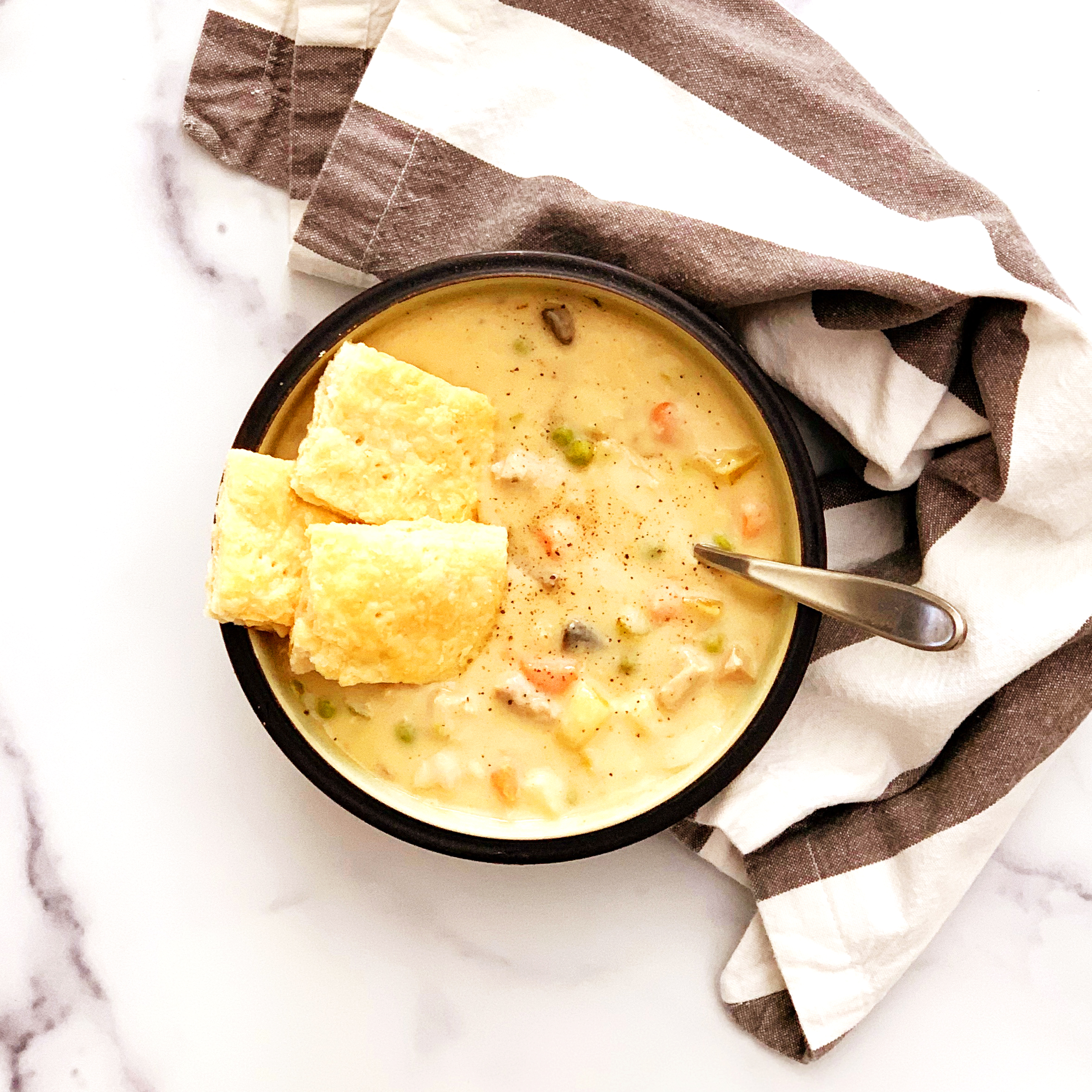 chicken pot pie soup in a bowl with pie crust crackers and a spoon in  bowl on a marble counter with a striped napkin.