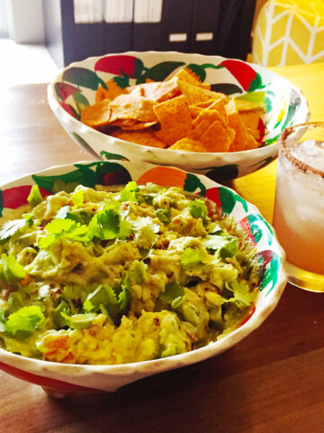 Guacamole in a bowl surrounded by chips and a margarita on a wood table
