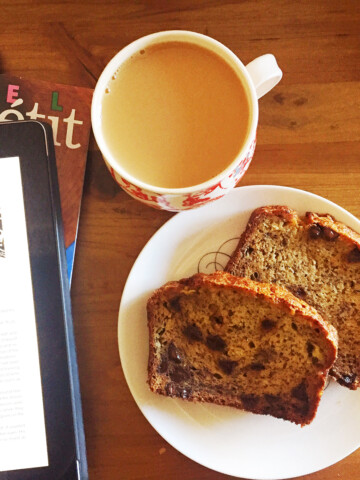 Chocolate chip banana bread on a white plate next to a cup of coffee and a kindle on a wood table