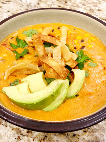 A bowl of chicken enchilada soup with avocado slices and tortilla strips on a counter.