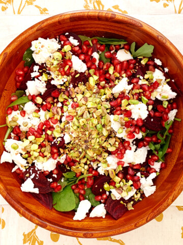 Beet, goat cheese, pomegranate and pistachio salad in a wood bowl on a white and gold tablecloth.