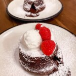 Two molten cholcolate lava cakes topped with powdered sugar, ice cream and raspberries on white plates on a wood tabletop