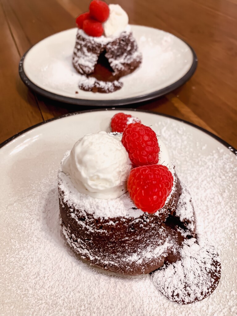 Two molten cholcolate lava cakes topped with powdered sugar, ice cream and raspberries on white plates on a wood tabletop