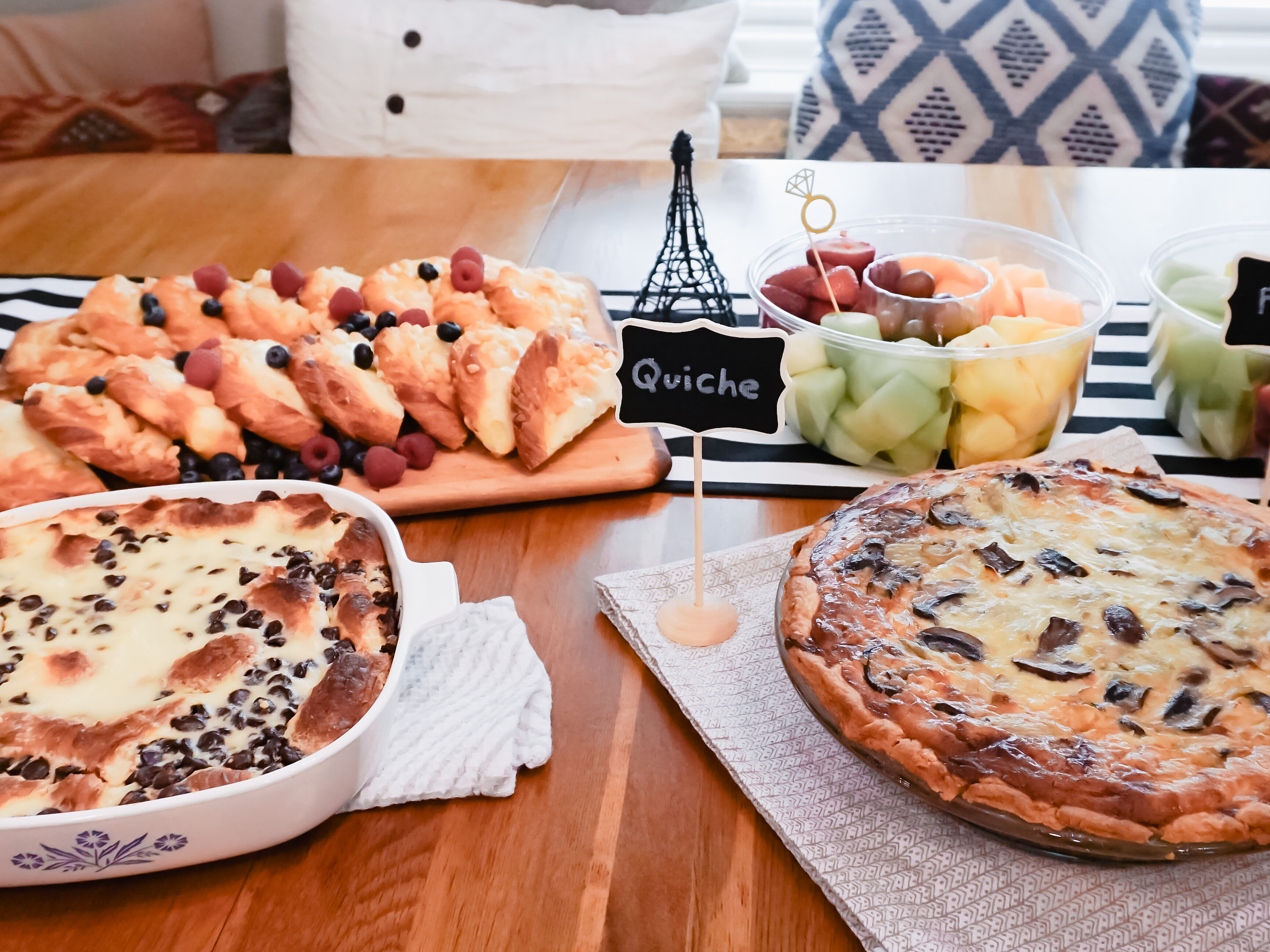 Chocolate croissant breakfast bake, danishes, quiche and fruit on a table with a small Eiffel Tower decoration in the back