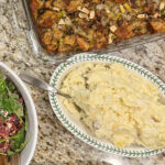 Roasted garlic mashed potatoes on a plate on a counter surrounded by salad and stuffing.
