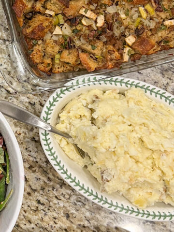 Roasted garlic mashed potatoes on a plate on a counter surrounded by salad and stuffing.