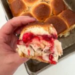 A turkey slider with cranberry sauce above a pan of turkey sliders.