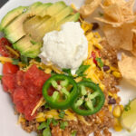 Mexican quinoa on a white plate with chips, salsa, avocado, Greek yogurt, cheese, and jalapeño slices.
